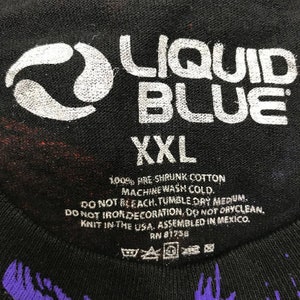 May include: Black t-shirt with a white Liquid Blue logo and a white swirl design. The shirt is size XXL and has care instructions printed on the tag. The tag says "100% Pre Shrunk Cotton, Machine Wash Cold, Do Not Bleach, Tumble Dry Medium, Do Not Iron Decoration, Do Not Dryclean, Knit in the USA, Assembled in Mexico, RN 81756."
