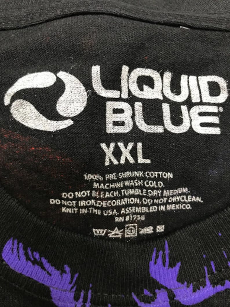 May include: Black t-shirt with a white Liquid Blue logo and a white swirl design. The shirt is size XXL and has care instructions printed on the tag. The tag says "100% Pre Shrunk Cotton, Machine Wash Cold, Do Not Bleach, Tumble Dry Medium, Do Not Iron Decoration, Do Not Dryclean, Knit in the USA, Assembled in Mexico, RN 81756."