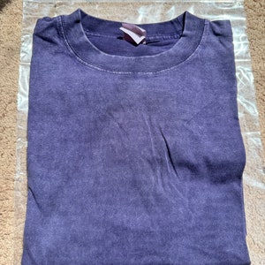 May include: A purple t-shirt with a faded look. The shirt is folded and placed on a light brown surface.