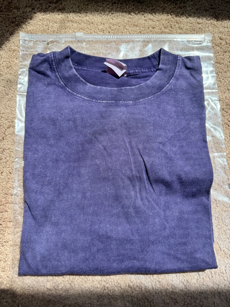 May include: A purple t-shirt with a faded look. The shirt is folded and placed on a light brown surface.
