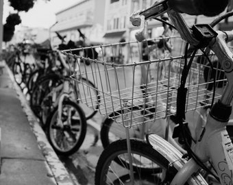 May include: A black and white photo of a row of bicycles parked on a city street. The bicycles have wire baskets and are lined up next to each other. The photo is taken from a low angle, looking up at the bicycles.