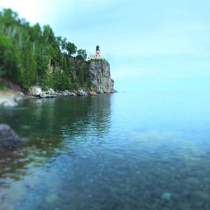 May include: A lighthouse perched on a rocky cliff overlooking a calm, clear blue lake. The water is shallow and the lakebed is visible. Lush green trees line the shore.