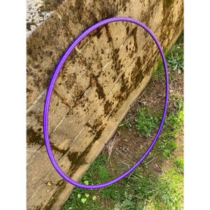 Peut inclure: Un cerceau d'exercice violet, &eacute;galement appel&eacute; hula hoop, est pos&eacute; sur le sol &agrave; c&ocirc;t&eacute; d'un mur en briques. Le cerceau est en plastique et a une surface lisse.