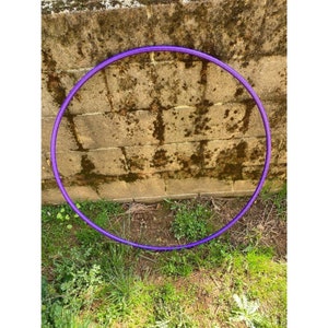 Peut inclure: Un cerceau d'exercice violet, &eacute;galement appel&eacute; hula hoop, est pos&eacute; sur le sol devant un mur en briques. Le cerceau est fait d'un mat&eacute;riau plastique flexible.