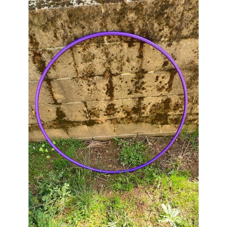 Peut inclure: Un cerceau d'exercice violet, &eacute;galement appel&eacute; hula hoop, est pos&eacute; sur le sol devant un mur en briques. Le cerceau est fait d'un mat&eacute;riau plastique flexible.