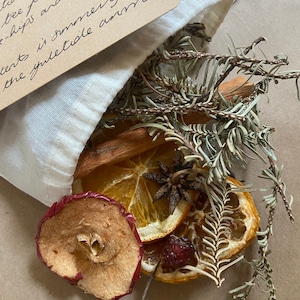 May include: A small, off-white fabric bag filled with dried botanicals. Visible are dried orange slices, cinnamon sticks, star anise, and pine sprigs. A brown tag with handwritten text is attached to the bag. The text on the tag reads "Yule Tide Simmer".