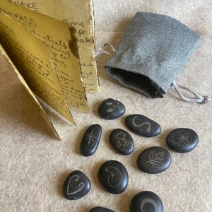May include: A set of 10 black stones with various symbols etched on them, including a goblet, a snake, a compass, a star, a leaf, a crescent moon, an eye, a heart, a swirl, and a flower. The stones are arranged on a beige surface next to a small gray pouch and a folded piece of paper with handwritten text.