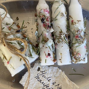 May include: White candles decorated with dried herbs, flowers, and gold flakes. One set is tied with twine. A handwritten tag reads "Yule & New Year manifestation candles."