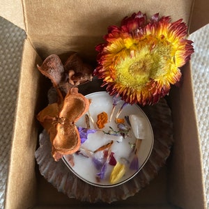 May include: A small, white candle with dried flowers and a clear crystal sits in a brown paper cup inside a cardboard box. A large, yellow and red flower and dried seed pods are also in the box.