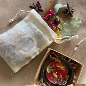 May include: A white muslin drawstring bag filled with dried herbs and spices, including cinnamon sticks, star anise, and dried berries. The bag is sitting on a brown paper surface.