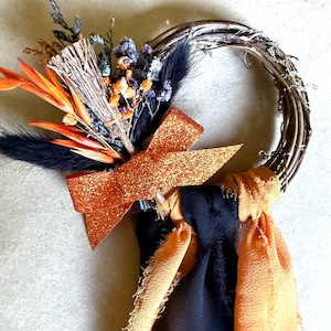 May include: A handmade autumn wreath with a grapevine base. It features orange and black accents, including a glittery orange bow, dried flowers, and black feathers. The wreath is approximately 10 inches in diameter.