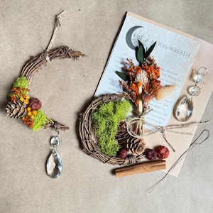 May include: Two decorative wreaths, one crescent-shaped and one circular, crafted from twigs and adorned with dried flowers, moss, and pinecones. A card with the text "MOON WREATH" and a crystal pendant are also present. The color palette includes natural browns, greens, and oranges.
