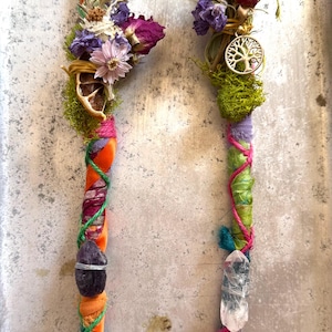 May include: Two handcrafted smudge sticks. Each stick features a wooden base wrapped with colorful fabric and twine, adorned with dried flowers, moss, and crystals. One has an amethyst, the other a clear quartz crystal, and a tree of life charm.