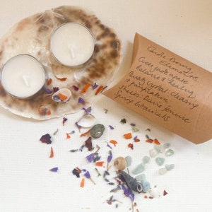 May include: A brown paper bag with the text "Candle Dreaming Elements. Green Jade Agate: balance & healing. Quartz Crystal: cleansing & purification. Shells: Divine feminine Spring botanicals" on it. Two white tea lights are on a seashell with purple and orange flower petals.