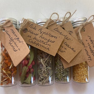 May include: Six small glass bottles with cork tops and twine tied around the necks. Each bottle contains dried herbs and spices. Each bottle has a small brown paper tag with handwritten text describing the contents. The text on the tags reads: "Marigold, Homegrown + Dried", "Pink Peppercorn, Hand Foraged in Southern California", "Lavender, Leaves + Flowers", "Rosemary", "Summer Savory, Foraged from the Woods + Mountain".