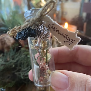 May include: A small glass vial filled with dried flowers and a small silver charm of a tree. The vial is tied with twine and a tag that reads "Grounding".