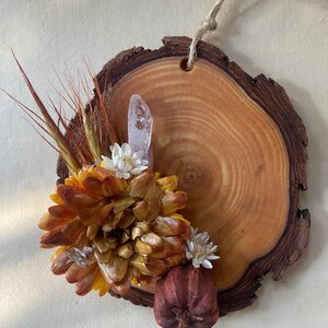 May include: A wooden slice ornament with a natural wood grain finish. It features a dried flower arrangement with a small, dried gourd and a clear crystal.