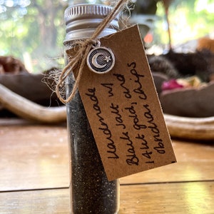 May include: A small glass bottle filled with black glitter and gold flakes. The bottle is tied with twine and a charm with a crescent moon and star. A brown paper tag is attached to the bottle with the text "Handmade Black Jade w/ 14kt gold & chips obsidian"