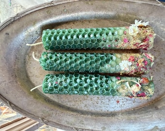 May include: Three green honeycomb-patterned candles with white wicks rest on a silver platter. The ends of the candles are decorated with gold glitter, dried flowers, and pink petals. The candles are a unique decorative item.