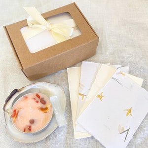 May include: A set of handmade paper stationery with pressed flowers and a small box containing a pink candle with dried flowers and a clear crystal.