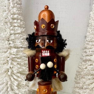 May include: A wooden nutcracker doll with a brown body, black hair, and a crown. The doll is wearing a white fur collar and white boots with gold accents. It is holding drumsticks and a small drum. The nutcracker is standing on an orange base.