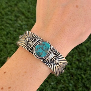 May include: Silver cuff bracelet featuring a large oval turquoise stone. The bracelet has a detailed design with sunburst patterns and decorative elements. The turquoise stone is a vibrant blue-green color, set in a silver bezel.