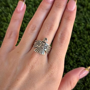 Op de afbeelding: Een zilverkleurige ring met een gestileerd vogelontwerp. De ring heeft een gedetailleerd, symmetrisch patroon met een centrale vogel figuur en geometrische accenten. De ring wordt aan een vinger gedragen, wat het ingewikkelde vakmanschap laat zien.