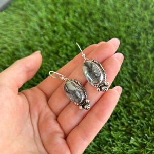 May include: A pair of silver earrings with oval-shaped green and black stones. The stones have a marbled pattern and are set in a silver frame with decorative details.