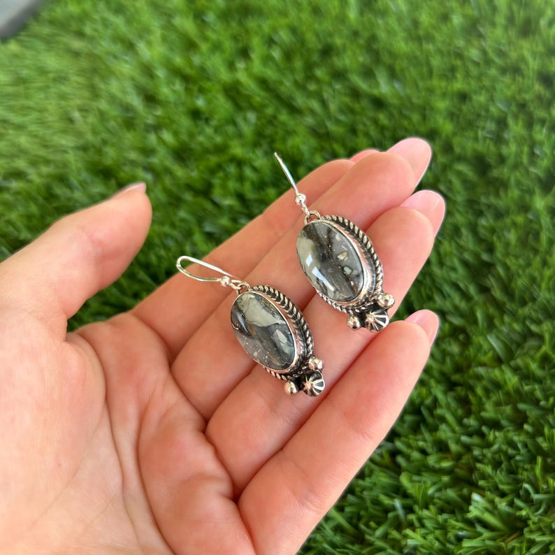 May include: A pair of silver earrings with oval-shaped green and black stones. The stones have a marbled pattern and are set in a silver frame with decorative details.