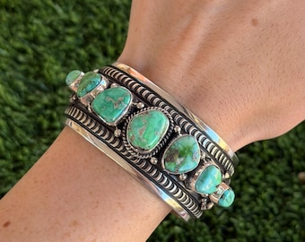May include: A wide silver cuff bracelet featuring several large, oval turquoise stones. The stones are a vibrant green-blue color and are set in ornate silver settings. The bracelet has a detailed, textured design.