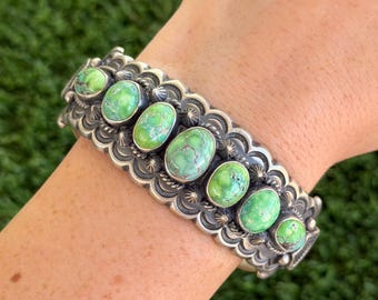 May include: A silver cuff bracelet featuring six oval-shaped green turquoise stones with brown matrix patterns. The metal band is decorated with intricate stamped scalloped edges, twisted wire detailing, and small silver bead accents.