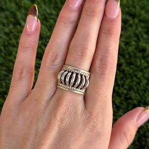 May include: A silver ring with a wide band featuring a series of vertical, rounded bars. The ring has a textured design with rope-like detailing along the top and bottom edges. The ring is worn on a finger.