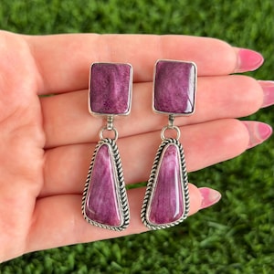 May include: A pair of sterling silver earrings featuring rectangular purple stones at the top and teardrop-shaped purple stones below. The teardrop stones are framed with a twisted silver rope design.