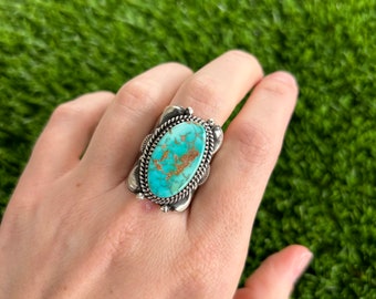 May include: A silver ring featuring a large oval turquoise stone set in a detailed silver bezel. The stone has a blue-green colour with brown matrix.