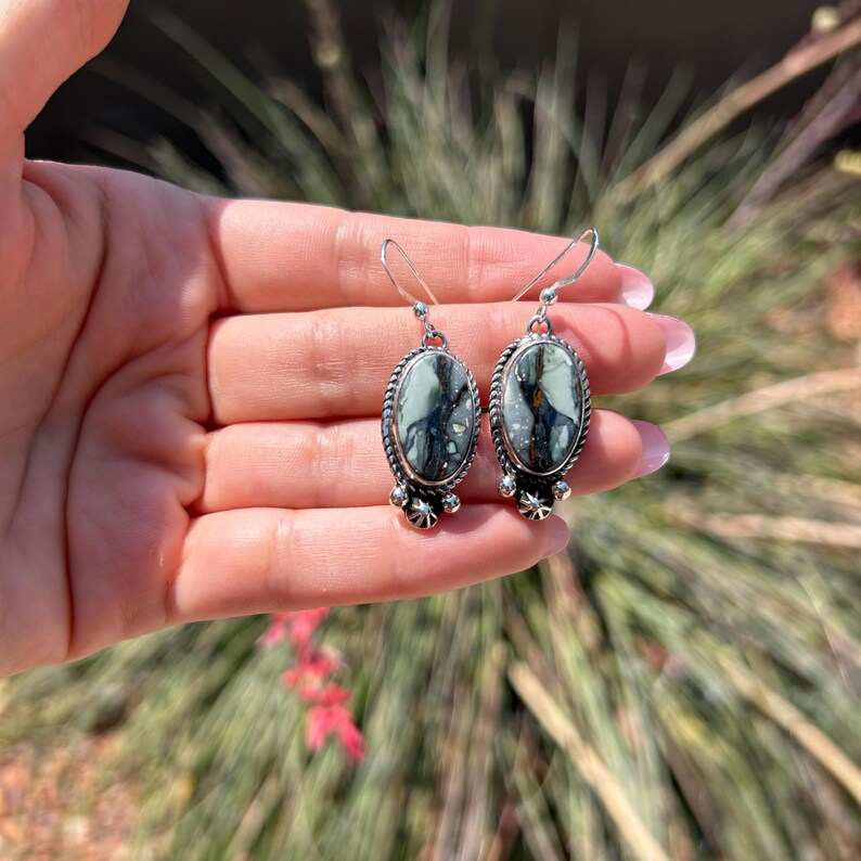 May include: A pair of silver dangle earrings with oval-shaped green and black gemstone stones. The earrings have a silver bead detail at the bottom.