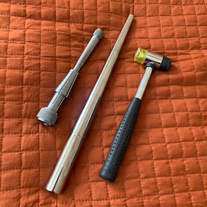 May include: Three metal tools for jewelry making: a silver ring mandrel, a silver ring sizing tool, and a black and yellow rubber mallet.