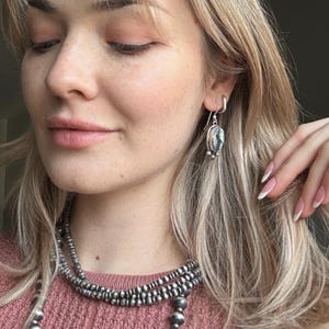 May include: A woman wearing a layered silver necklace and silver earrings with a dark gray stone. The earrings are dangle earrings with a small silver bead at the bottom.