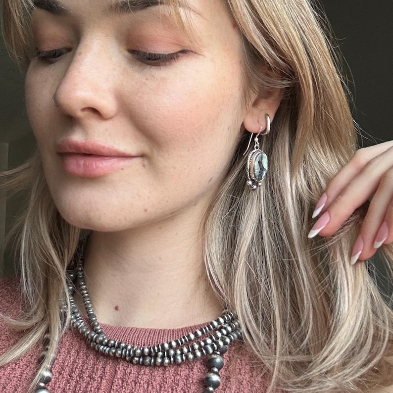 May include: A woman wearing a layered silver necklace and silver earrings with a dark gray stone. The earrings are dangle earrings with a small silver bead at the bottom.