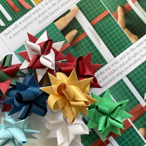 Pode incluir: Uma coleção de oito estrelas de papel em várias cores, incluindo vermelho, branco, azul, verde e dourado. As estrelas são dobradas de tiras de papel e estão dispostas em um tapete de corte verde.