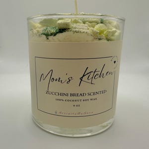 May include: A clear glass candle jar with a zucchini bread scented candle. The candle has a white label with the text "Mom's Kitchen" and "Zucchini Bread Scented". The candle is 9 oz and made with 100% coconut soy wax.