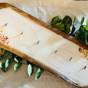 Pode incluir: Uma vela em bandeja de madeira com uma base de cera branca e flores secas incrustadas na cera. A bandeja &eacute; oval e tem um acabamento de madeira natural.