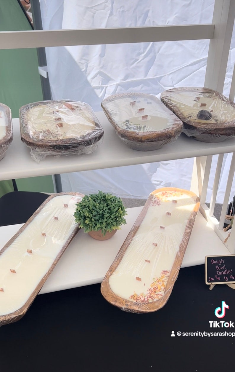 Pode incluir: Uma exibi&ccedil;&atilde;o de velas de tigela de massa de madeira. As velas s&atilde;o brancas e t&ecirc;m flores secas embutidas. As velas est&atilde;o em uma prateleira branca com uma placa que diz "Velas de tigela de massa 3 in - $18 1g - $14".
