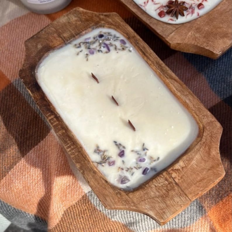 Pode incluir: Uma vela em uma tigela de madeira com uma base de cera branca e detalhes em roxo e branco. A vela est&aacute; sobre um cobertor xadrez.