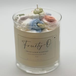 May include: A clear glass candle jar filled with off-white wax, topped with white, blue, pink, green, and orange decorative elements. The label reads "Fruity-O" and "Fruit Loop Scented". The candle is made with 100% coconut soy wax and is 9 oz.