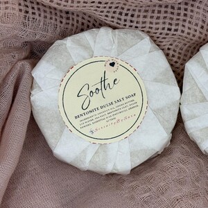 May include: Round, wrapped soap with a cream-colored label that reads "Soothe" in cursive. The label also lists ingredients like coconut oil and sea salt. The soap is wrapped in white paper and sits on a pink fabric background.