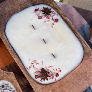 Pode incluir: Uma vela branca com detalhes vermelhos e marrons em uma tigela de madeira. A vela tem tr&ecirc;s pavios e &eacute; decorada com anis estrelado e flocos vermelhos.