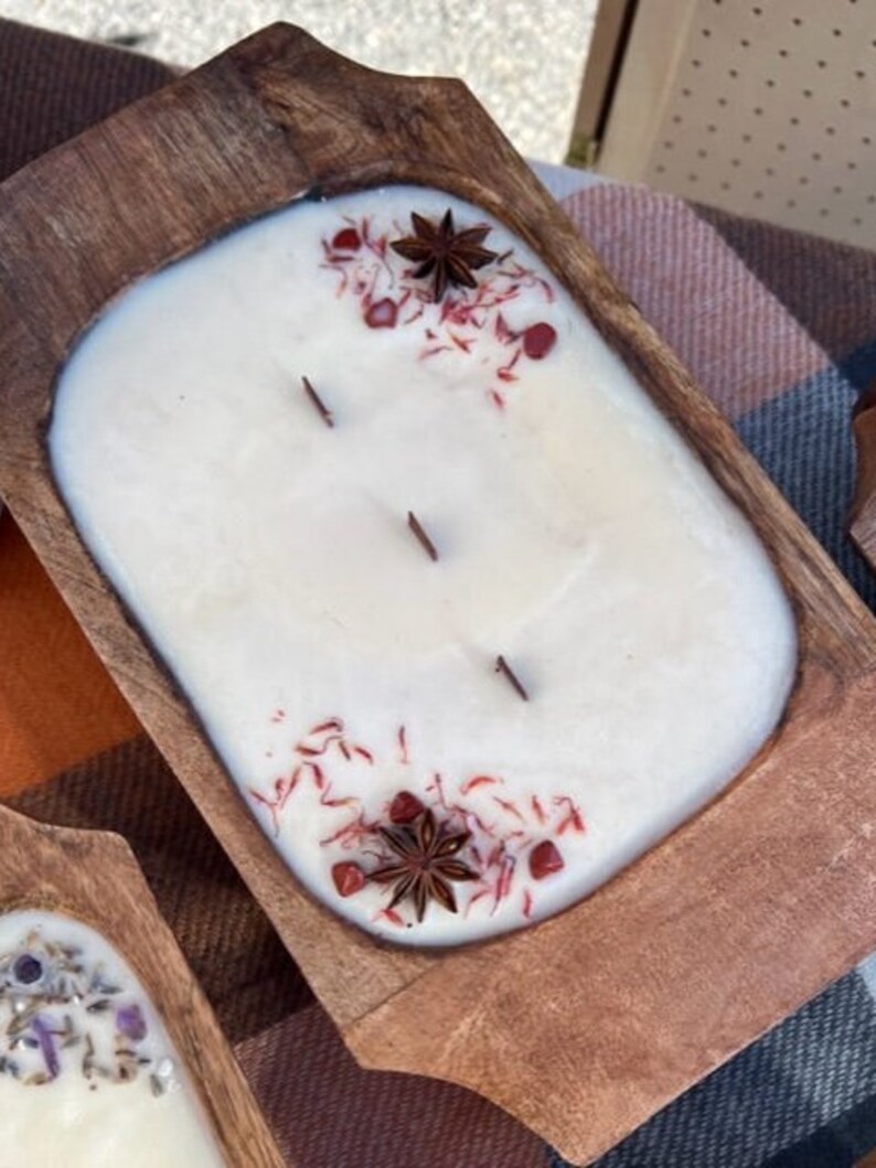 Pode incluir: Uma vela branca com detalhes vermelhos e marrons em uma tigela de madeira. A vela tem tr&ecirc;s pavios e &eacute; decorada com anis estrelado e flocos vermelhos.