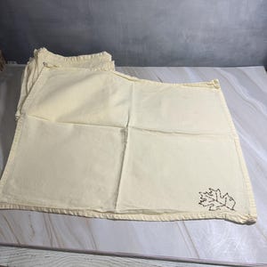 Op de afbeelding: Een stapel cr&egrave;mekleurige placemats. Elke placemat heeft een gestikt bruin bladerontwerp in de hoek. De placemats zijn rechthoekig met een gevouwen rand.