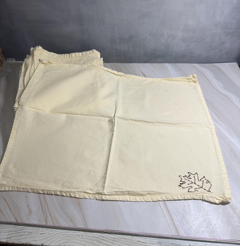 Op de afbeelding: Een stapel cr&egrave;mekleurige placemats. Elke placemat heeft een gestikt bruin bladerontwerp in de hoek. De placemats zijn rechthoekig met een gevouwen rand.