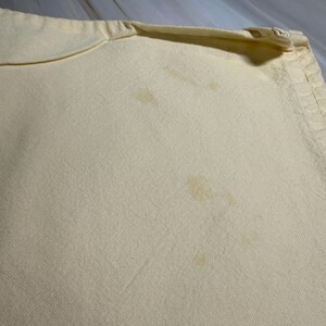 Op de afbeelding: Close-up van een lichtgele kussensloop met zichtbare vlekken. De kussensloop heeft een ritssluiting aan &eacute;&eacute;n kant en een licht gebogen rand. De stof lijkt een geweven materiaal te zijn.